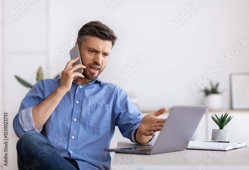 Obraz na płótnie Angry millennial businessman talking on mobile phone and looking at laptop scree