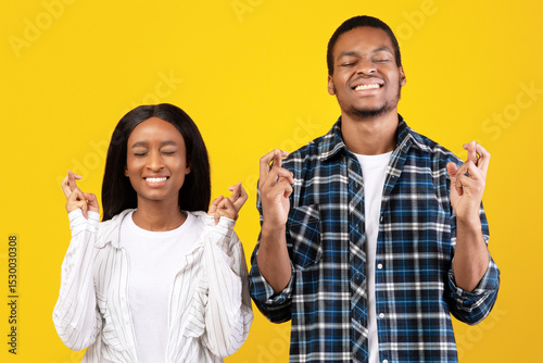 Make a wish, really want a dream come true. Excited millennial african american students man and lady in casual with fingers crossed and closed eyes, isolated on yellow background, empty space photo