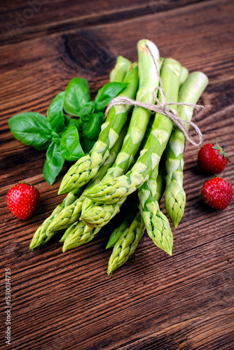 Bunch raw green asparagus offered as close-up on a rustic wooden board with fresh strawberries