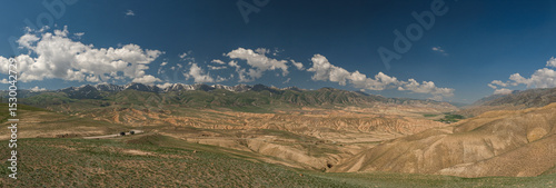 Wide angle landscape panorama of a winding road going through a highland mountain pass, high peaks with glaciers in the background. Summer day, blue sky with white clouds. Kyrgyzstan, Naryn Region.