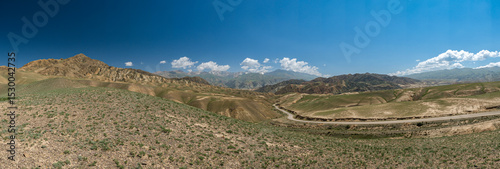 Wide angle landscape panorama of a winding road going through a highland mountain pass, high peaks with glaciers in the background. Summer day, blue sky with white clouds. Kyrgyzstan, Naryn Region.