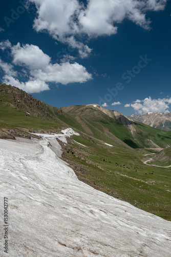 Wide angle landscape of a winding road going through a highland mountain pass surrounded by high peaks with glaciers. Summer day, blue sky with white clouds. Kyrgyzstan, Naryn Region, Central Asia.