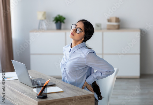 Tired upset young indian business woman suffering from back pain, muscle spasm at workplace with laptop in office or home interior. Fatigue, deadline, pain and incorrect posture, work at computer