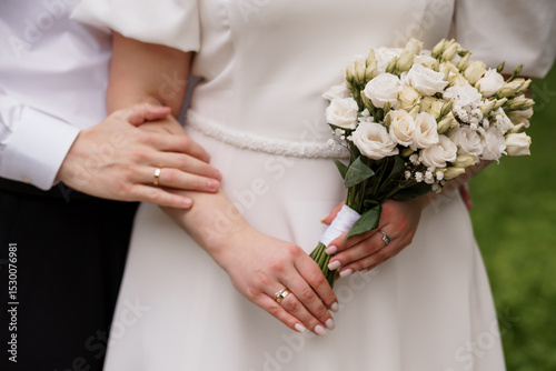 Romantic Wedding Day Close-Up: Bride and Groom Hands with Rings, White Bouquet, Elegant Dress, Outdoor Greenery, Symbolic Love and Commitment.