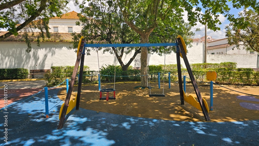 Fototapeta premium Empty swings in shaded playground surrounded by trees — childhood memories and peaceful atmosphere
