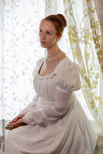 A Regency period woman wearing a white embroidered cotton dress and surrounded by backlit lace curtains