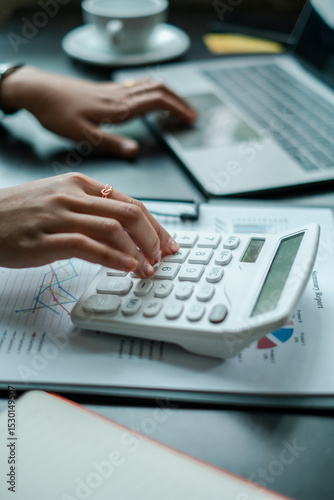 Close-up of hands using a calculator and laptop, analyzing financial data in a modern office environment.