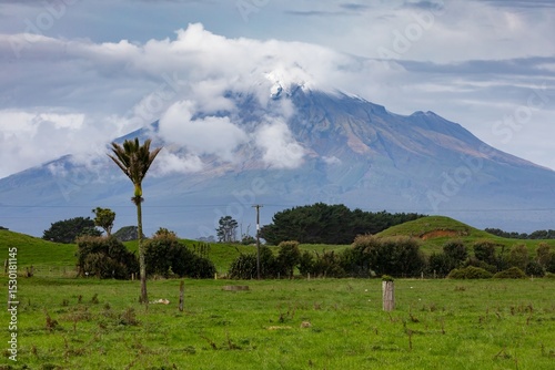 Mount Taranaki, a dormant volcano, rises above green fields in Pungarehu, Taranaki, New Zealand. Clouds obscure the peak, creating a dramatic landscape.