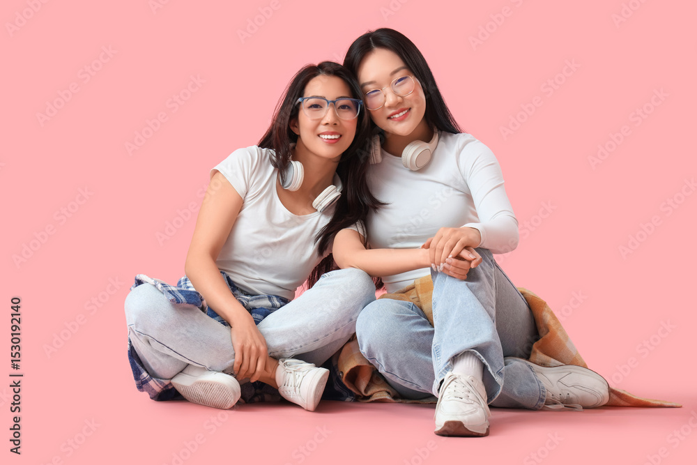 Beautiful young happy Asian sisters with headphones sitting on pink background Beautiful young happy Asian sisters with headphones sitting on pink background