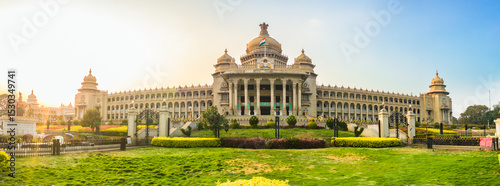 Panoramic view of Vidhana Soudha, the iconic government building in Bengaluru, India, captured during golden hour. The lush gardens and clear sky frame the historic architecture beautifully