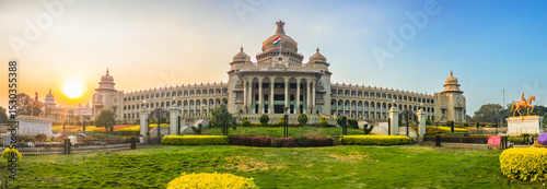 Panoramic view of Vidhana Soudha, the iconic government building in Bengaluru, India, captured during golden hour. The lush gardens and clear sky frame the historic architecture beautifully
