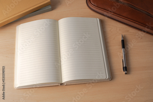 Top-down view of an open blank lined notebook on a light wooden desk, with a black and silver pen, a leather case, and journal. perfect for themes of journaling, planning, education, or office work.
