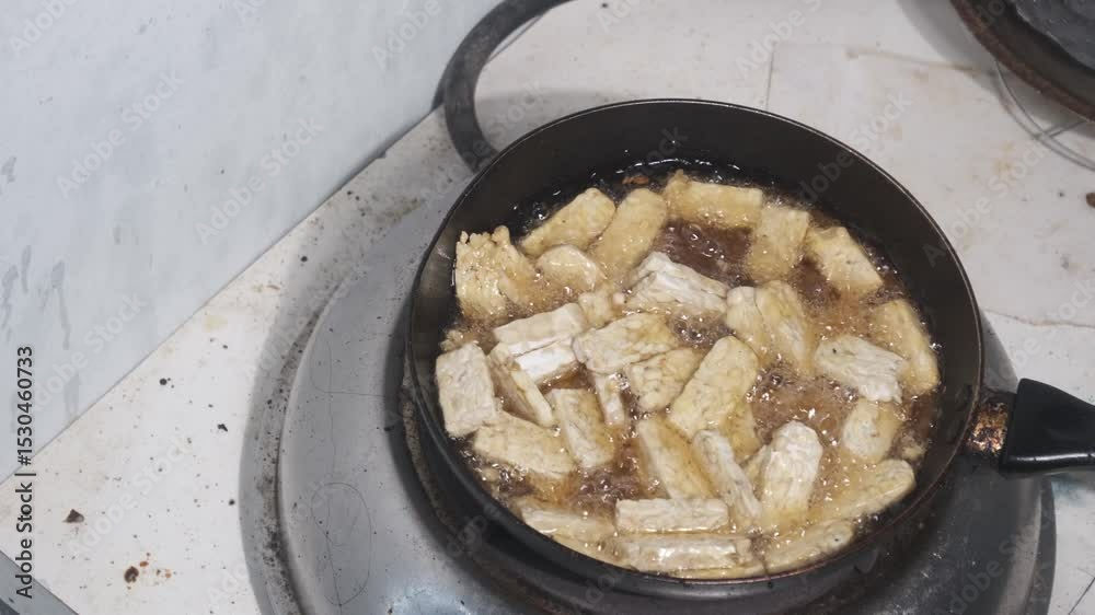 Raw tempeh is being fried in bubbling hot oil, with fresh white slices sizzling in the pan. The intense heat creates foam and motion around the pieces as they begin to cook. This close-up scene. 4K