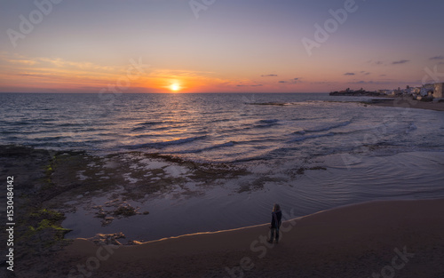 Woman Gazes at the Vibrant Sunset Over the Tranquil Sea in Punta Secca, Sicily, Italy