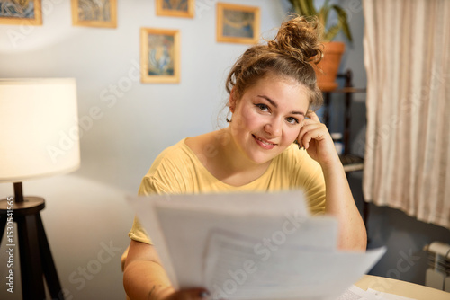Horizontal image of beautiful smiling caucasian female with hair knob holding paper docs in hand sitting at table in her cozy living-room, feeling happy after receiving money refund or loan approval