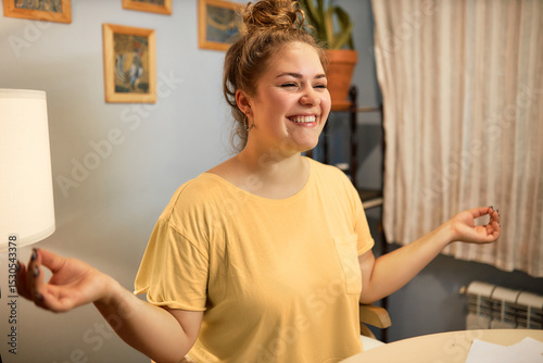 Side view of smiling cheerful female in yellow t-shirt having rest or break doing yoga or meditating to relieve stress and relax making mudra gesture with hands while working online from home