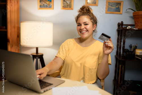 Happy woman consumer spending her money on shopping online holding credit card and using laptop, smiling at camera while doing purchase on marketplace with big discount , buying clothes