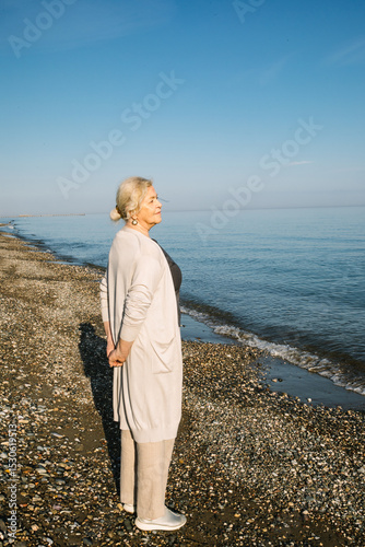 Εκτύπωση καμβά Elderly woman stands on a quiet beach with hands behind her back, looking at the sea
