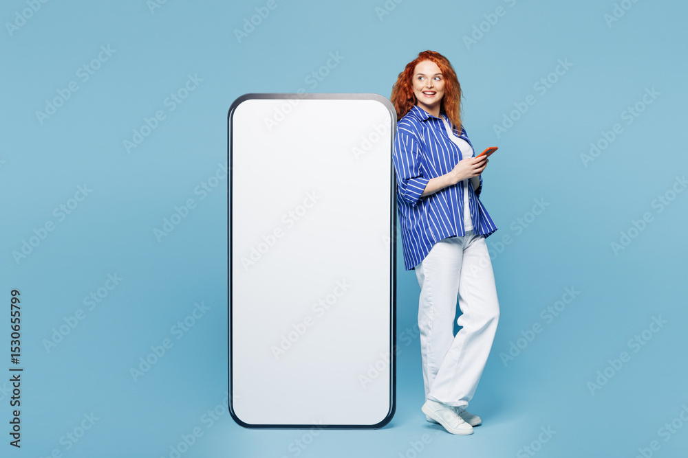 Full body side view young ginger woman wear shirt t-shirt casual clothes stand big huge blank screen mobile cell phone with workspace area use smartphone isolated on plain light blue cyan background. Full body side view young ginger woman wear shirt t-shirt casual clothes stand big huge blank screen mobile cell phone with workspace area use smartphone isolated on plain light blue cyan background.