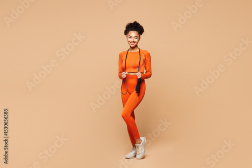Full body young happy fitness trainer instructor woman sportsman she wear orange clothes spend time in gym jumping on skipping rope isolated on plain beige background. Workout sport fit abs concept.