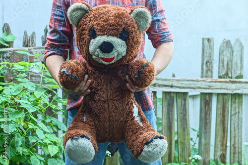 man in checkered shirt and jeans holding in hands one big old toy soft plush brown baby gift with black eyes and nose and white ears and face torn teddy bear outside