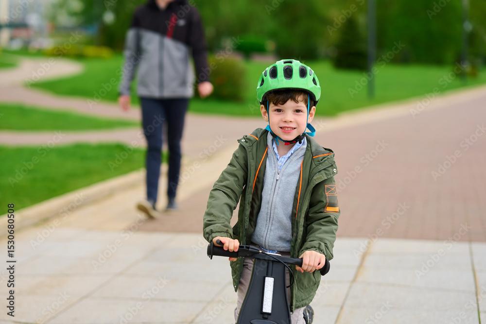 Obraz premium child riding scooter in park, wearing helmet outdoors. smiling boy enjoying leisure time on sunny day, embodying joy and safety. parent in background simulates supervision, relaxation
