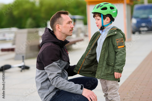 father instructing boy in green helmet on sidewalk next to scooter, emphasizing safety and bonding outdoors. focus on protection, parenting, and quality family time in fresh air