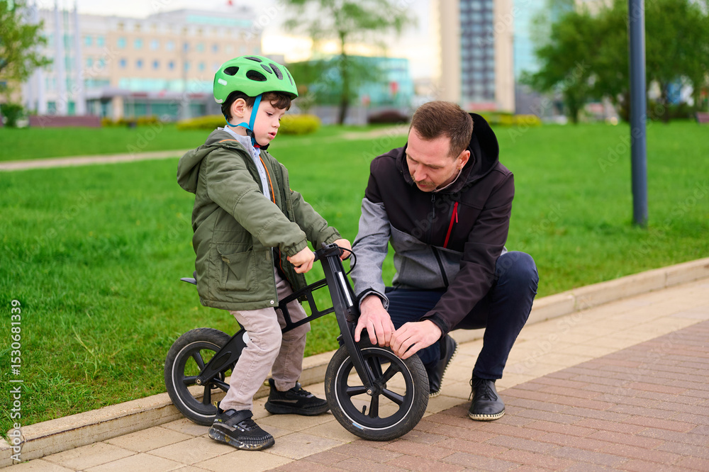 Fototapeta premium father teaching son balance bike riding in park. learning safety, outdoor activity, focus on bonding and guidance. urban background, fatherhood and child's development theme