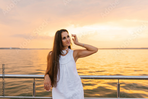 Young woman in white dress stands on pier overlooking the ocean. The sky is a mix of orange and pink hues, creating a serene and peaceful atmosphere