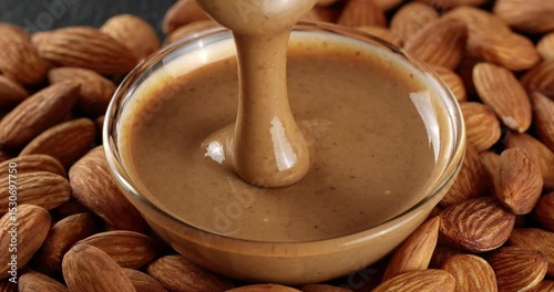 Almond nut butter is being scooped with a spoon. The almond butter drips from the spoon into a glass bowl on a background of almond nuts