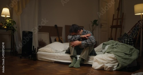 Side view of a sad male artist in a checkered shirt sitting on a white mattress and crying into his hands after a hard and desperate day. Artists difficult experiences