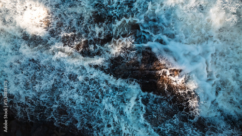Aerial top-down view of powerful ocean waves crashing over coastal rocks, creating abstract white foam patterns. Perfect for backgrounds, marine themes, nature textures, and environmental projects.