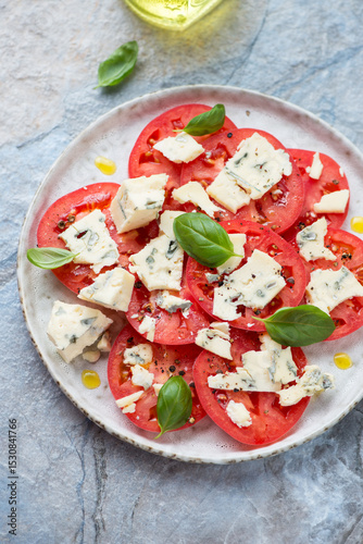 Sliced tomatoes with blue cheese and green basil, vertical shot on a blue and beige stone background, elevated view