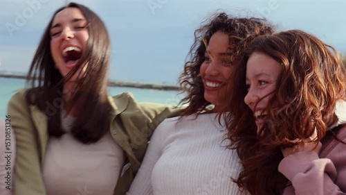 Female friends from diverse backgrounds walking, laughing, and hugging along sandy shoreline, sharing carefree moments during sunny summer vacation