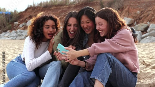 Four cheerful young women, likely students or recent graduates, are enjoying their time together on a sandy beach, capturing memories with their smartphone