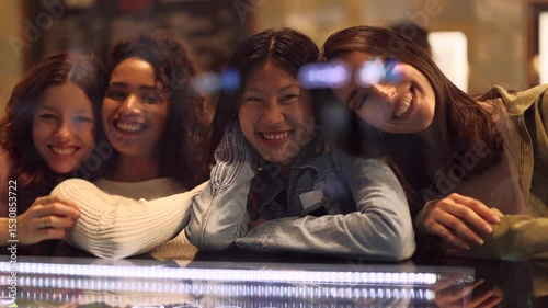 Young women sharing playful moment, making faces behind pastry shop window, expressing joy and camaraderie during lighthearted group interaction