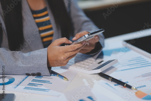 Attractive and cheerful millennial Asian businesswoman receiving a good message or email through her smartphone, sitting at her desk.