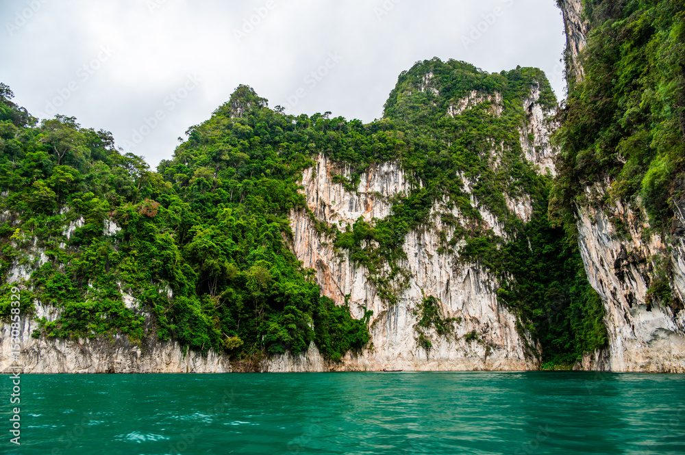 View of Khao Sok Lake in Krabi province, Thailand, with towering limestone cliffs covered in tropical rainforest rising above the calm surface of the lake. View of Khao Sok Lake in Krabi province, Thailand, with towering limestone cliffs covered in tropical rainforest rising above the calm surface of the lake.