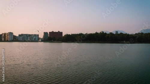 Forward drone footage starting over Tirana’s artificial lake at sunset, rising to reveal the city skyline framed by trees and distant mountains in the capital of Albania.