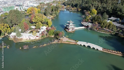 A serene view of Black Dragon Pool in Lijiang, with calm waters reflecting ancient architecture. Snow-capped mountains rise in the distance. Trees sway gently in the breeze. A peaceful moment in natur