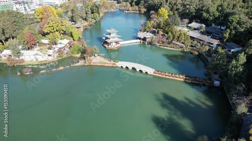 A serene view of Black Dragon Pool in Lijiang, with calm waters reflecting ancient architecture. Snow-capped mountains rise in the distance. Trees sway gently in the breeze. A peaceful moment in natur