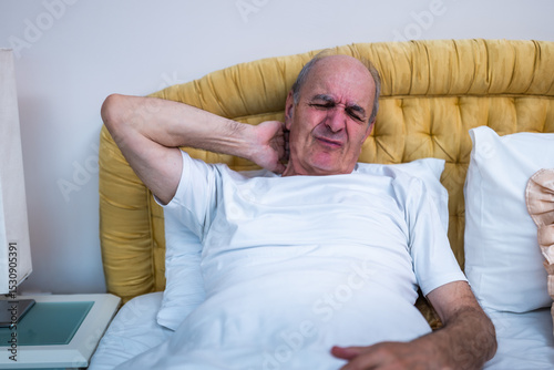 Worried senior man suffering from neck pain and discomfort after waking up in bed.