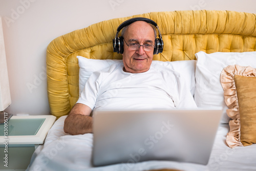 Happy elderly man in pajamas using  laptop and wearing headphones while sitting in bed.