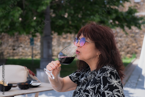 Senior woman wearing sunglasses drinking a glass of red wine at an outdoor restaurant in summer