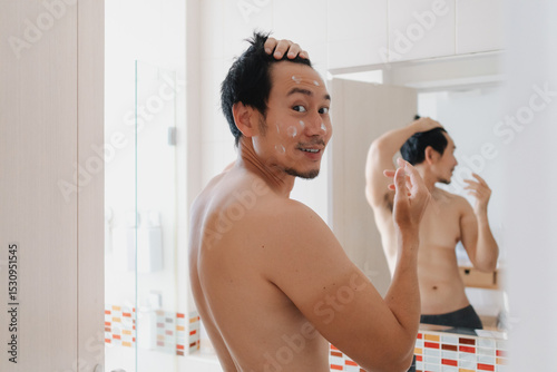 Smiling Thai man applying cream in bathroom mirror