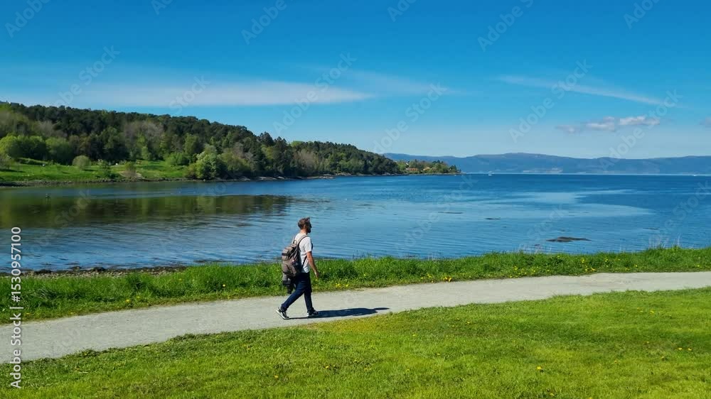 A young man strolls along the shoreline in Lade by a Norwegian fjord on a sunny spring day, enjoying the stunning landscape and clear blue sk