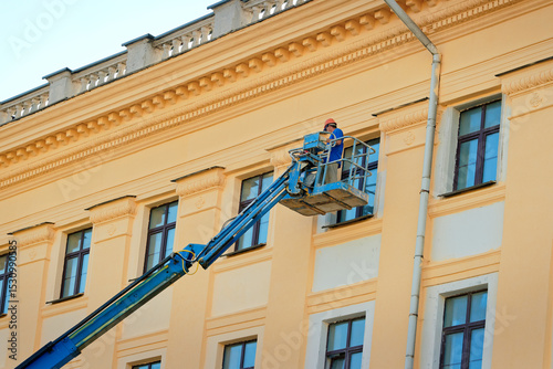 Worker on an elevated boom lift repairing and painting facade of building, routine maintenance and exterior renovation. Construction worker on mobile lift working on an ornate building facade