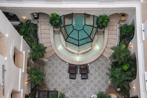Canvas-taulu Interior of a boutique hotel spa featuring a glass enclosed pool, curved stone pathway, lush greenery, patterned tiles, and elegant white arches