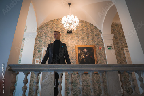 Elegant hotel interior with a man on a stone balustrade, ornate wallpaper, chandelier, classical portrait, arched doorways, and decorative columns.