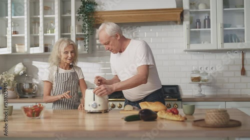 Aged man and woman trying new toaster in home kitchen, portrait of old spouses. Housewife and her husband viewing and examining new kitchen appliance for cooking breakfast, aged man cutting bread